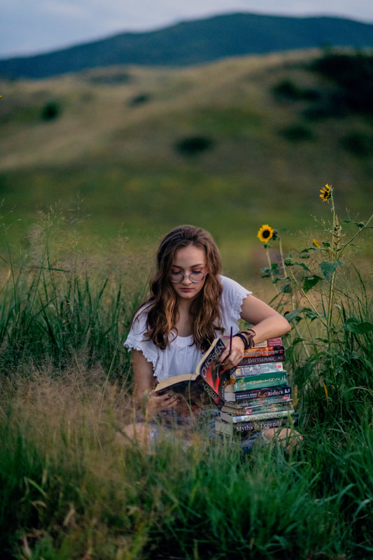 Girl in white shirt, jeans, and glasses surrounded by books in a field in golden colorado senior pictures outside